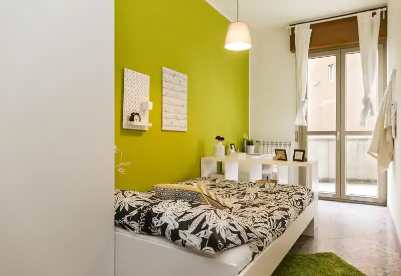 Camera da letto moderna e luminosa con giaciglio bianco e copripiumino decorato, parete accent verde e una scrivania vicino alla finestra che lascia entrare molta luce naturale.