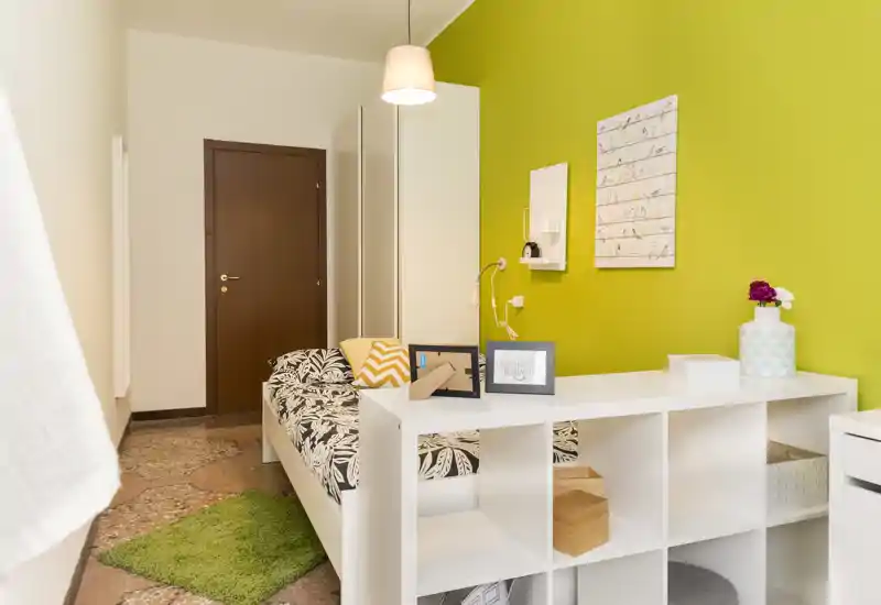 Camera singola moderna e luminosa con letto con motivo, libreria/box a piedi del letto e parete accentuata verde — ideale per un affitto compatto e di design.