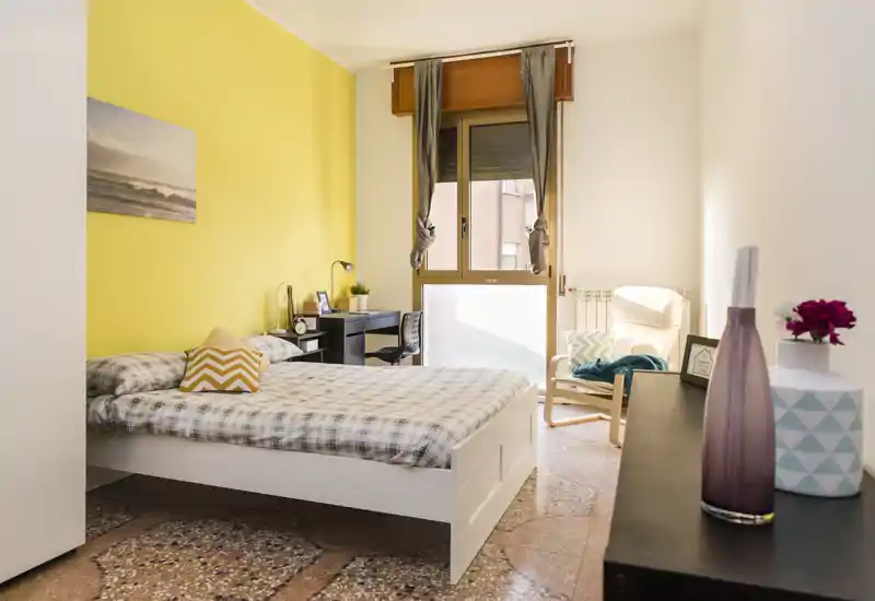 Camera da letto luminosa e curata con letto matrimoniale, scrivania vicino alla finestra e comoda poltrona; parete gialla e luce naturale rendono l'ambiente accogliente per gli annunci.