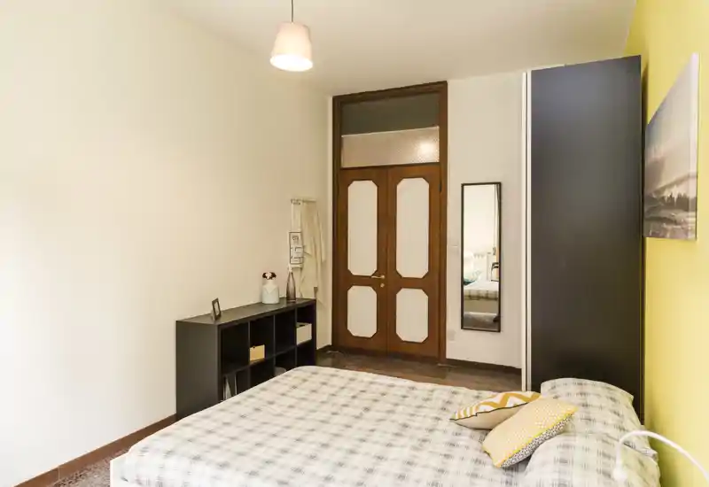 Camera da letto luminosa e ben allestita con un letto matrimoniale in primo piano, cuscini decorativi, un armadio alto, una libreria a cubi e uno specchio a figura intera vicino alla porta.