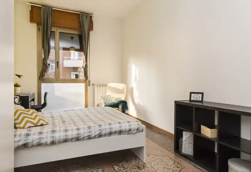 Camera da letto luminosa e arredata in modo semplice con letto matrimoniale, biancheria a motivo, poltrona vicino alla finestra e una libreria bassa — luce naturale calda.