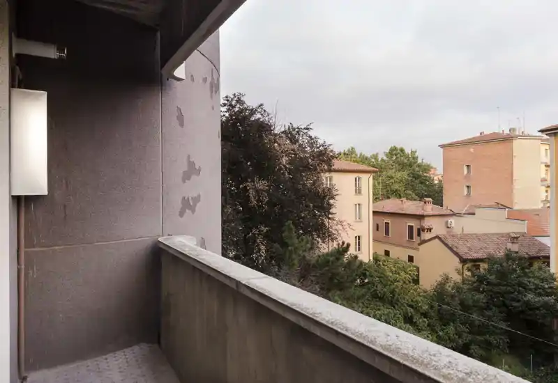 Small private balcony with concrete balustrade overlooking neighboring buildings and trees; soft natural light and urban view, suitable for morning coffee.