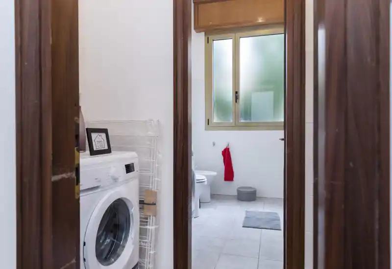 Compact bathroom with tiled floor, frosted window and simple fixtures; a washing machine sits just outside the doorway and a red towel adds a pop of color.