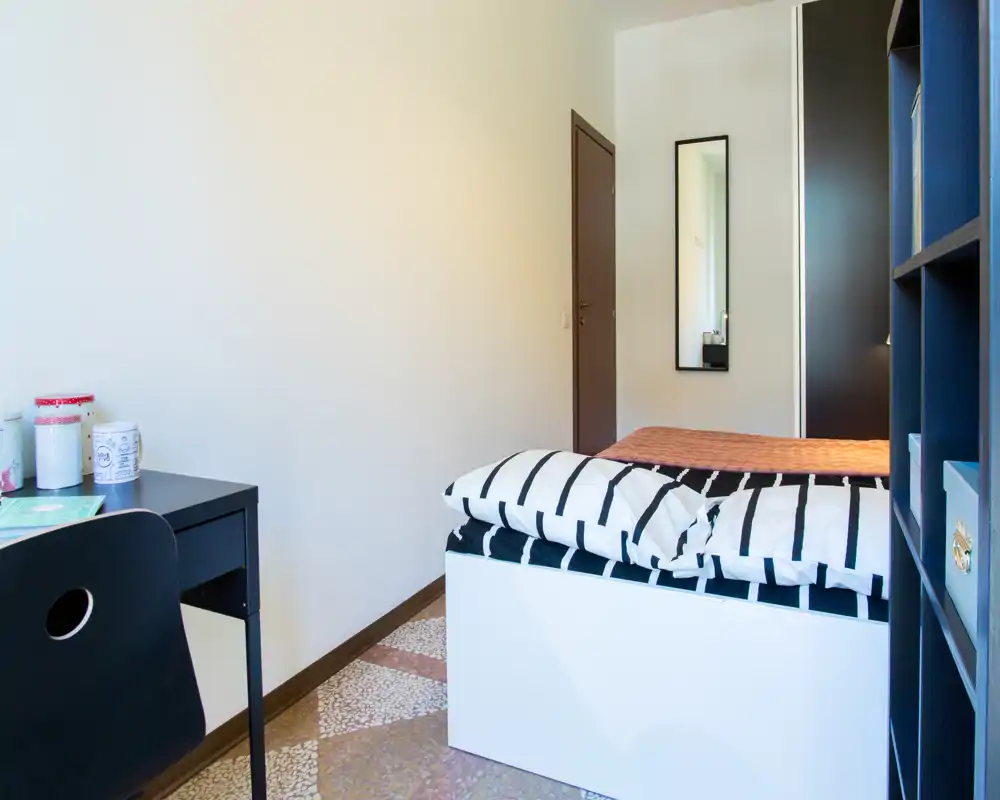 Camera da letto compatta e luminosa con letto singolo ben rifatto, una scrivania e scaffalatura per riporre gli effetti personali — stile minimal pulito, adatta a una persona.
