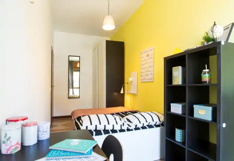 Camera da letto singola luminosa e ordinata con copriletto a righe, libreria a cubi e armadio davanti a una parete gialla vivace.