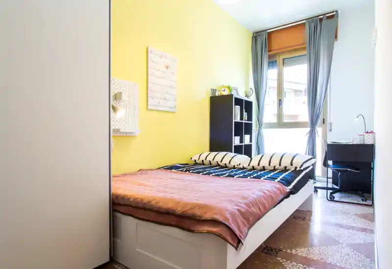 Camera da letto luminosa e ben arredata con letto matrimoniale, parete gialla e luce naturale dalla finestra. Mensole semplici e una piccola scrivania rendono lo spazio funzionale e accogliente, ideale per un annuncio.