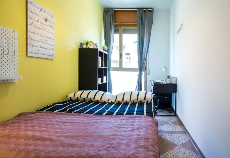 Camera da letto luminosa e ben arredata con letto matrimoniale, biancheria a righe, finestra con tende e una piccola scrivania — accogliente e adatta a brevi soggiorni.