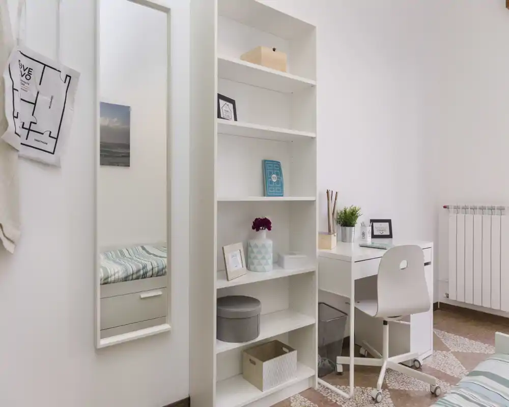 Coin de chambre lumineux et soigné avec lit visible, miroir mural, étagère et petit bureau blanc — style minimaliste et touches décoratives agréables.