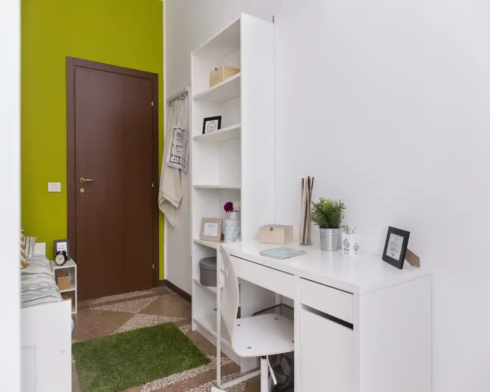 Coin chambre lumineux et rangé avec un bureau blanc, des étagères et un lit simple partiellement visible près d'un mur vert. L'espace de travail est décoré de plantes et de cadres, idéal pour une location compacte ou une chambre étudiante.