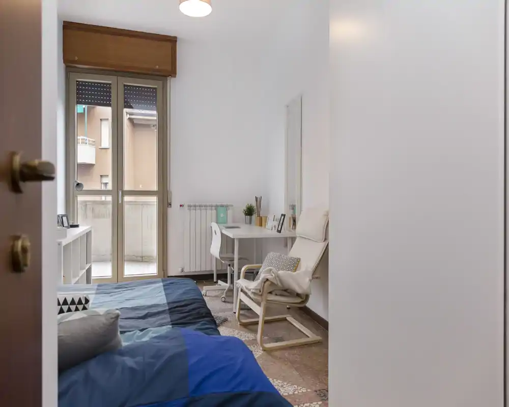 Camera da letto luminosa e ordinata con letto matrimoniale, scrivania e porta finestra verso il balcone che garantisce luce naturale. Arredo neutro e una poltrona accogliente creano un ambiente tranquillo.