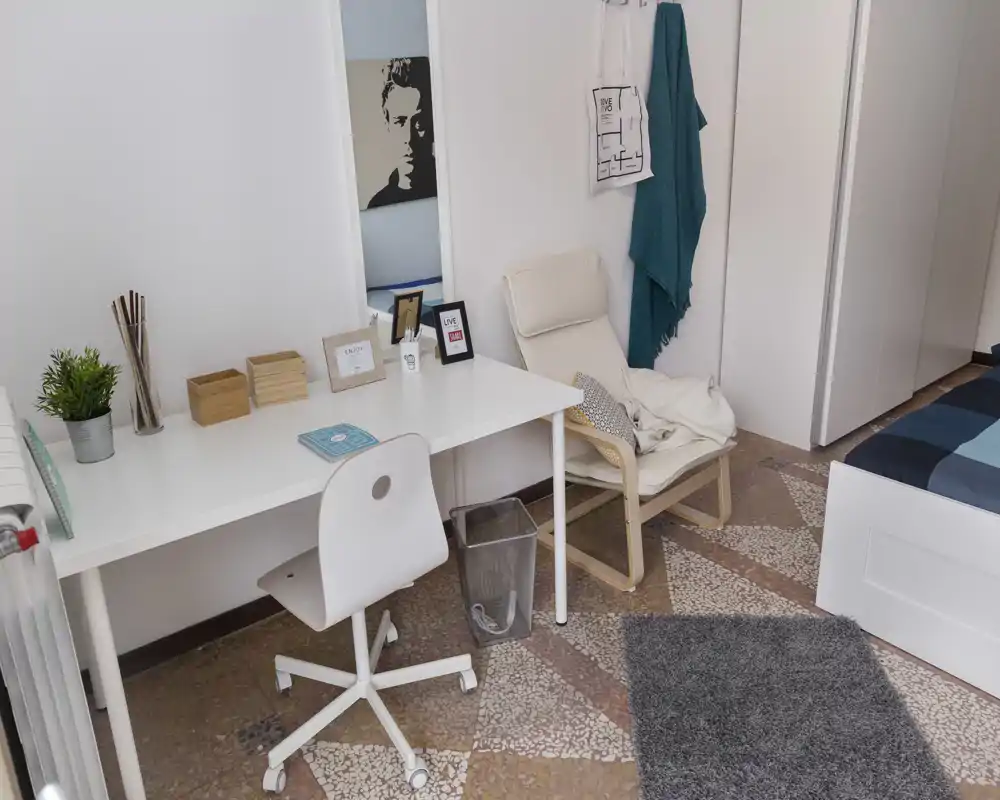 Angolo camera luminoso e ordinato con scrivania, poltrona e parte del letto. Ideale per uno studente o single, lo spazio offre un'area studio e tessili confortevoli.