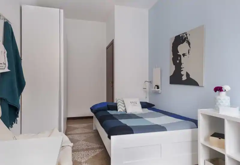 Camera da letto singola ben arredata con letto rifatto, parete azzurra, armadio e mensole — ambiente luminoso e accogliente ideale per un singolo.