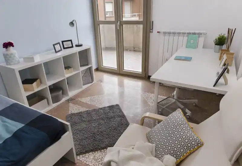 Camera da letto moderna e luminosa con letto singolo, scrivania e scaffalatura vicino a una porta finestra che dà su un piccolo balcone; arredo ordinato con cuscini e tappeto.