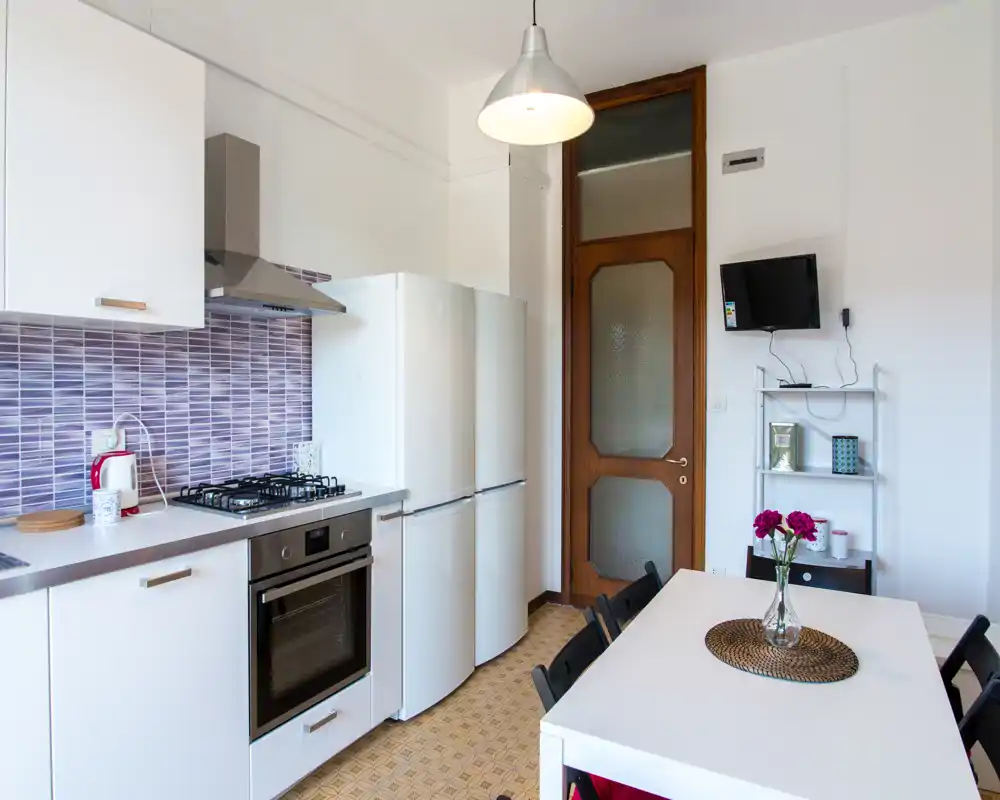 Cucina moderna e luminosa con mobili bianchi, forno integrato, piano cottura a gas e frigorifero. Un tavolo da pranzo con sedie e fiori freschi crea un ambiente accogliente e ben curato, perfetto per annunci.
