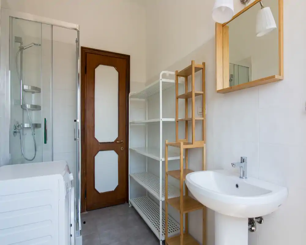 Bagno pulito e luminoso con box doccia in vetro, lavabo a colonna, scaffalature e zona lavatrice; piastrelle neutre e dettagli in legno donano un aspetto moderno e funzionale.