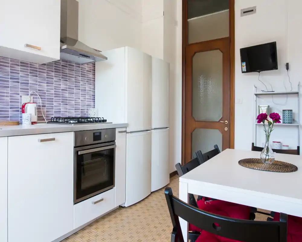 Cucina ben attrezzata con mobili bianchi, forno integrato, piano cottura a gas e frigorifero a colonna. Un tavolo da pranzo con sedie e fiori freschi crea un angolo accogliente dove mangiare.