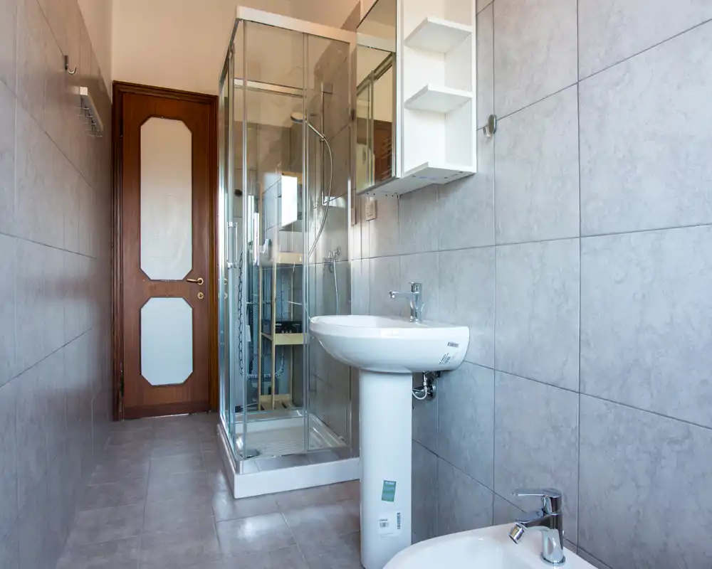 Bagno moderno e luminoso con piastrelle a parete, lavabo a colonna, bidet e cabina doccia in vetro. Toni neutri e sanitari puliti creano un ambiente funzionale.