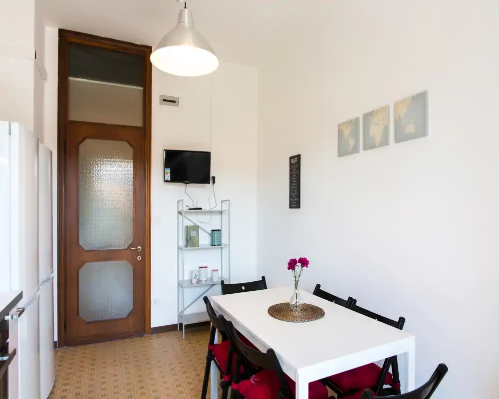 Zona pranzo luminosa e ordinata con un tavolo bianco, sedie pieghevoli nere con cuscini rossi e un semplice centrotavola floreale — ideale per pranzi informali.
