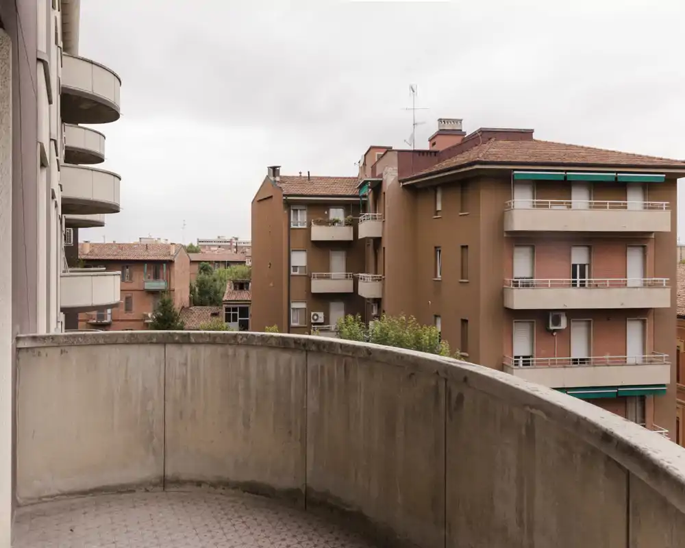 Piccolo balcone curvo che si affaccia su edifici residenziali; parapetto in cemento e pavimento piastrellato con cielo nuvoloso, ideale per descrivere uno spazio esterno urbano.