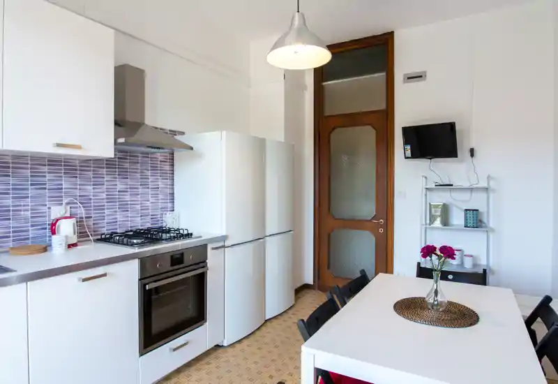 Bright modern kitchen with white cabinets, built-in oven, gas hob and a refrigerator. A dining table with chairs and fresh flowers creates a welcoming, well-styled space ideal for rental listings.