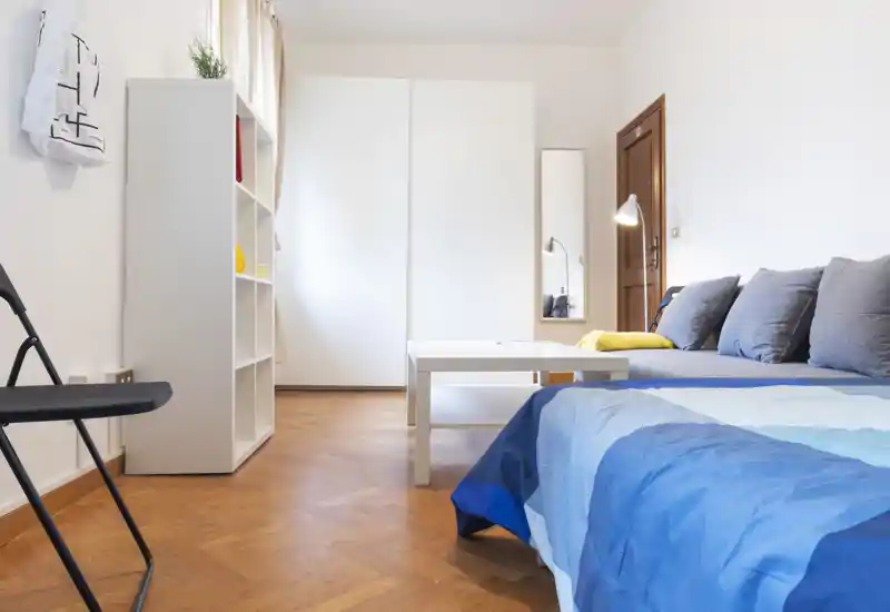 Dormitorio moderno y luminoso con colcha azul, cojines grises y una mesa de centro compacta; unidad de almacenamiento blanca y armario que aportan un estilo minimalista.
