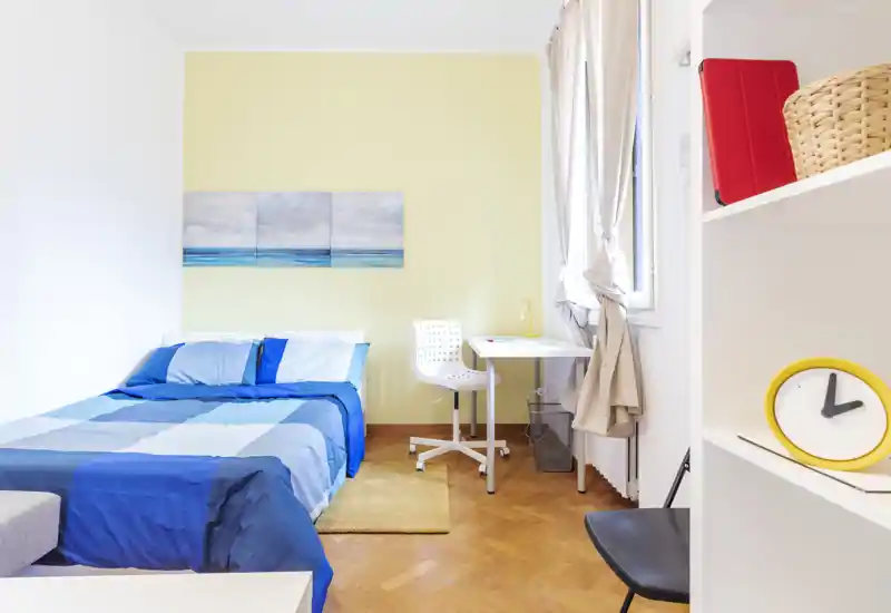 Dormitorio luminoso y bien presentado con cama doble de ropa de cama azul, pequeño escritorio junto a la ventana y suelos de madera cálidos — ideal para una estancia acogedora.