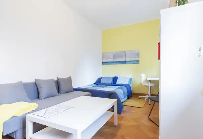 Camera da letto luminosa e ben arredata con letto matrimoniale in biancheria blu, divano e tavolino in primo piano — perfetta per un monolocale compatto.