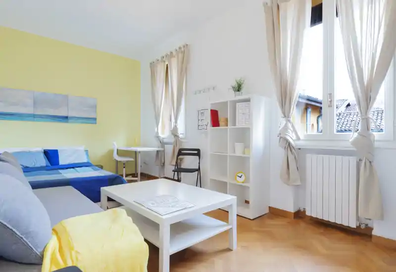 Dormitorio luminoso y acogedor con cama de rayas azules, pequeño espacio de estar y grandes ventanas que aportan mucha luz natural. Decoración neutra, estantería blanca y suelo de parquet que crean un ambiente agradable para alquileres.