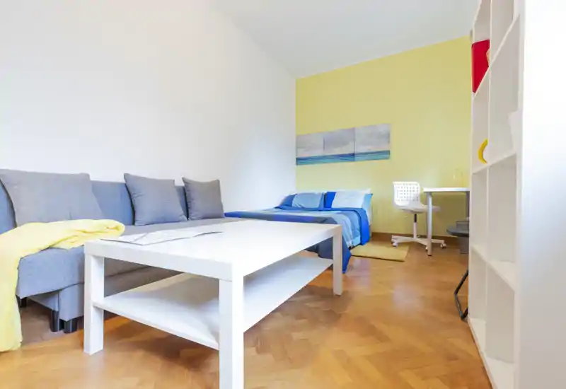 Dormitorio tipo estudio luminoso y bien presentado con cama azul, sofá y mesa de centro blanca. Suelo de parquet y detalles suaves crean un ambiente acogedor, perfecto para un anuncio de alquiler.