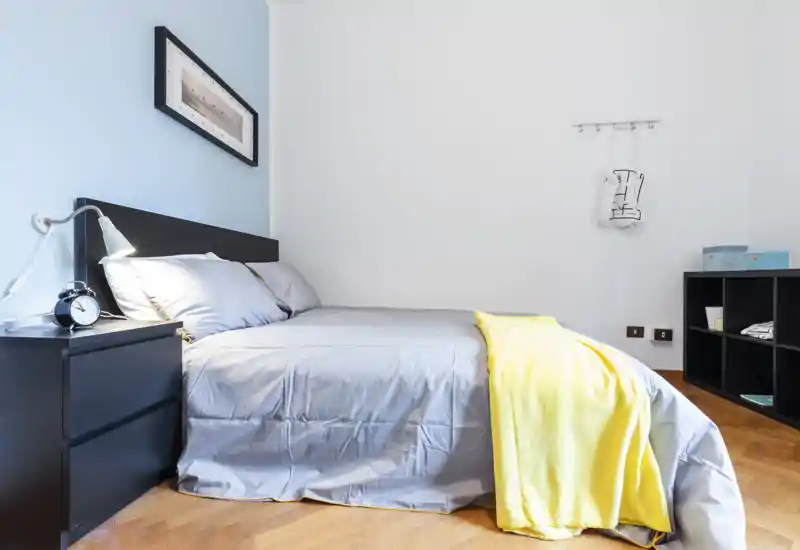 Dormitorio moderno y luminoso con cama doble bien hecha, ropa de cama gris y una manta amarilla. Líneas limpias, suelo de madera y almacenamiento minimalista crean un espacio tranquilo y acogedor para alquileres.