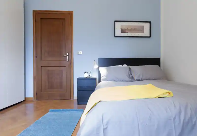Dormitorio luminoso y ordenado con cama doble vestida en ropa de cama gris y manta amarilla, mesita con lámpara y puerta de madera: habitación moderna y acogedora ideal para anuncios.