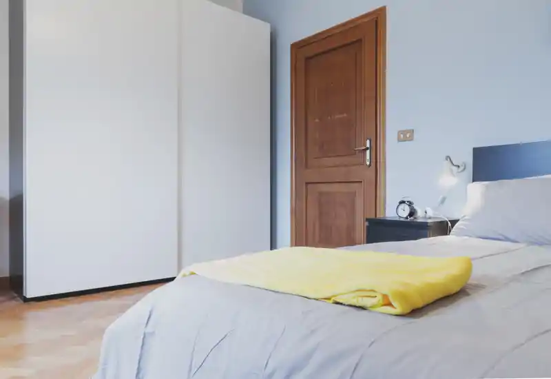 Dormitorio luminoso y ordenado con cama doble cómoda, mesita con lámpara y despertador, y un gran armario blanco — ideal para anuncios que destaquen un espacio para dormir moderno y limpio.
