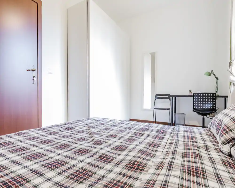 Dormitorio luminoso y minimalista con una cama grande en primer plano, armario y un pequeño escritorio junto a la ventana — ideal para estancias cómodas y teletrabajo.