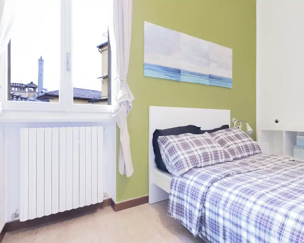 Dormitorio luminoso y ordenado con cama doble vestida con ropa de cuadros, cabecero blanco y cuadro en la pared. La gran ventana aporta luz natural que hace la habitación aireada y acogedora.