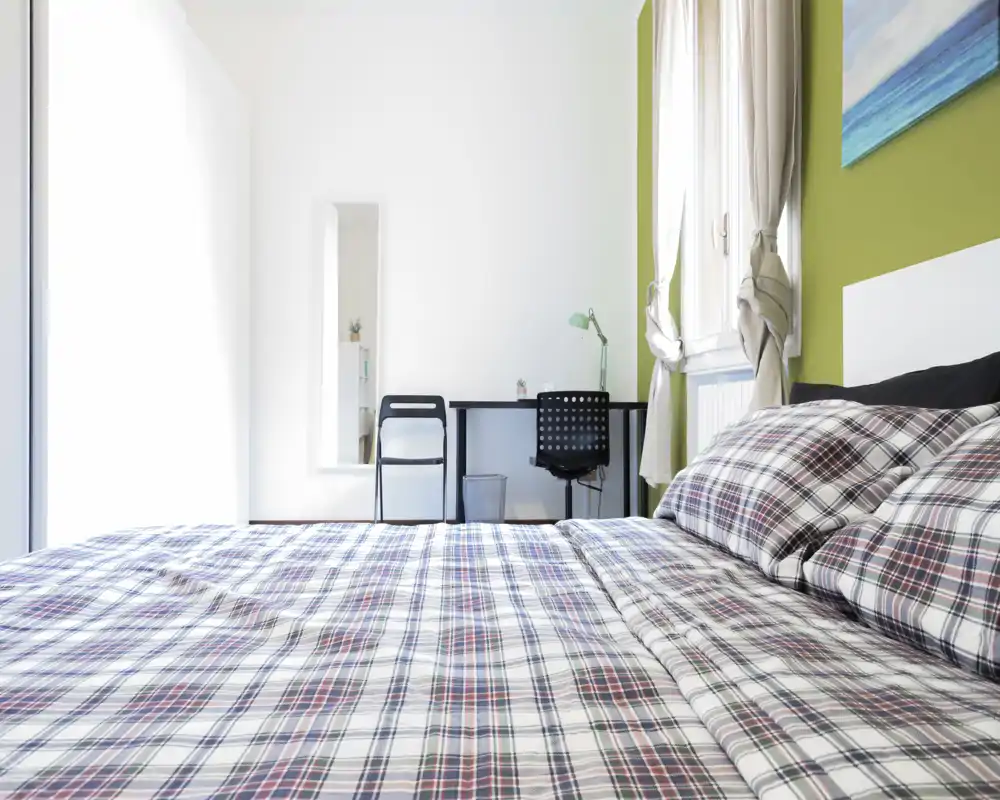 Dormitorio moderno y luminoso con una gran cama estampada en primer plano, un pequeño escritorio junto a la ventana y luz natural abundante.