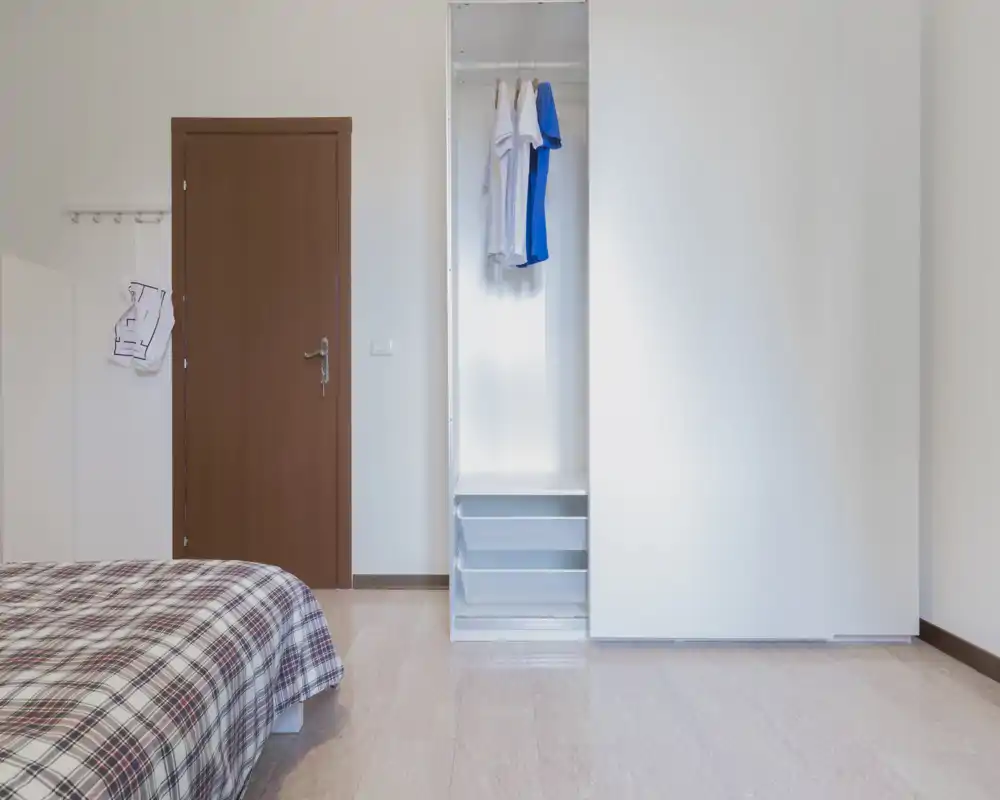 Rincón de dormitorio luminoso y minimalista con cama parcialmente visible con edredón de cuadros y armario empotrado blanco con puertas correderas; decoración neutra y líneas limpias para un aspecto moderno.