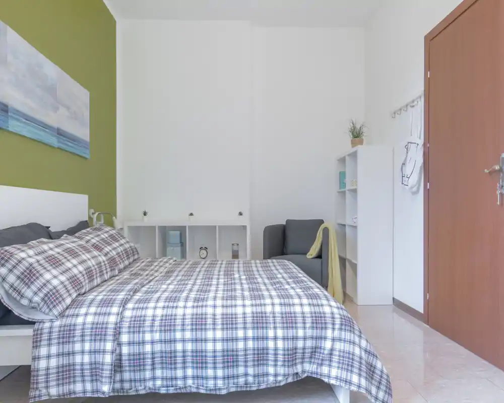 Dormitorio luminoso y bien presentado con una cama grande a cuadros como punto focal, acompañado de un sillón compacto y estanterías para almacenamiento — ideal para anuncios de alquiler.