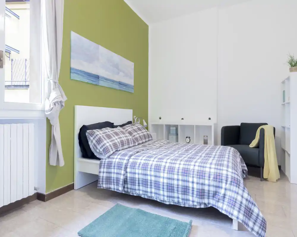 Dormitorio luminoso y ordenado con cama doble cómoda, ropa de cama a cuadros y luz natural desde la ventana. Un sillón y estanterías minimalistas completan el ambiente moderno y acogedor.