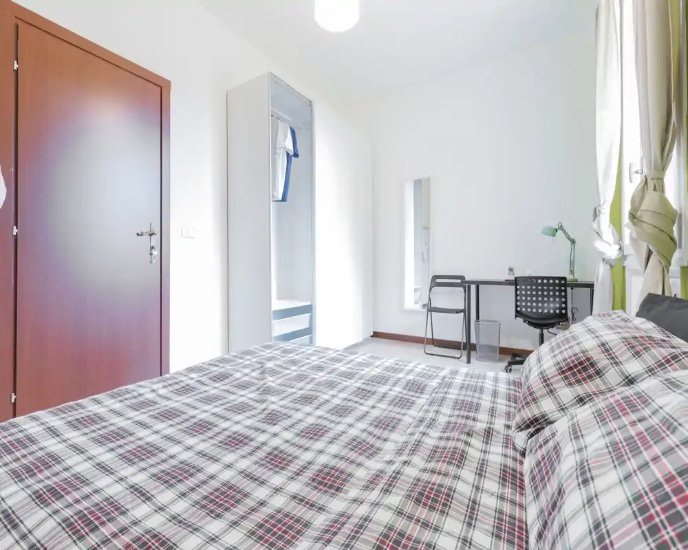 Dormitorio moderno y luminoso con una gran cama en primer plano, armario, escritorio y ventana con cortinas que aportan luz natural.