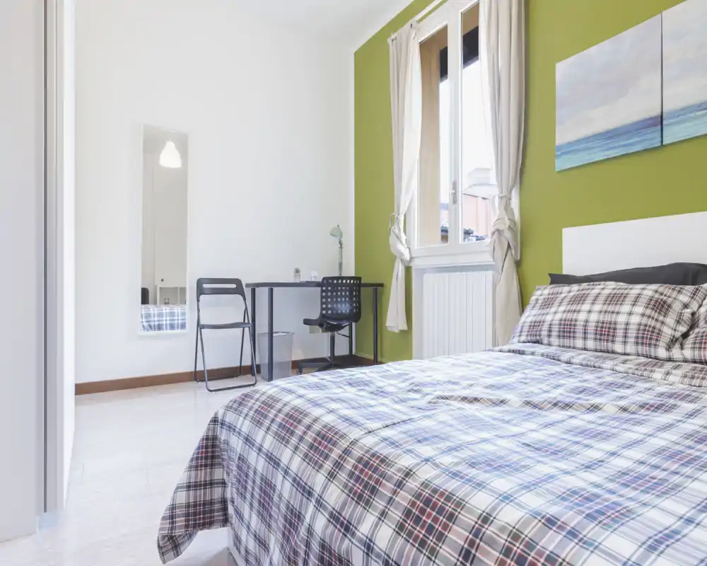 Dormitorio luminoso y ordenado con cama grande vestida con ropa de cuadros, un escritorio y luz natural desde la ventana; pared verde y decoración sencilla que aportan calidez.