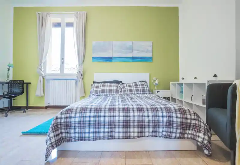 Dormitorio luminoso y bien presentado con una amplia cama doble, ropa de cama a cuadros y un tríptico con motivos marinos sobre la cabecera. La luz natural de la ventana aporta una atmósfera fresca y acogedora, ideal para anuncios de alquiler.