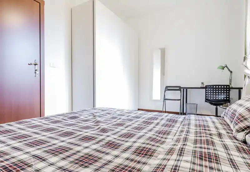 Dormitorio luminoso y minimalista con una cama grande en primer plano, armario y un pequeño escritorio junto a la ventana — ideal para estancias cómodas y teletrabajo.