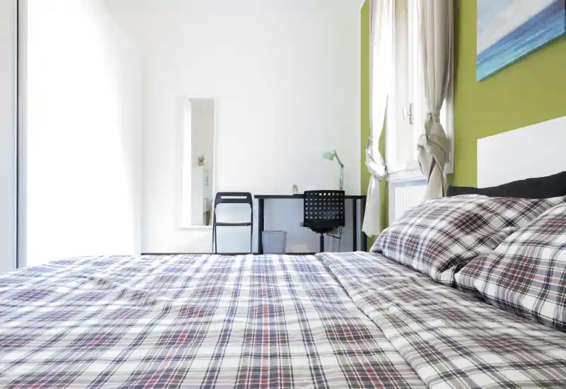 Dormitorio moderno y luminoso con una gran cama estampada en primer plano, un pequeño escritorio junto a la ventana y luz natural abundante.