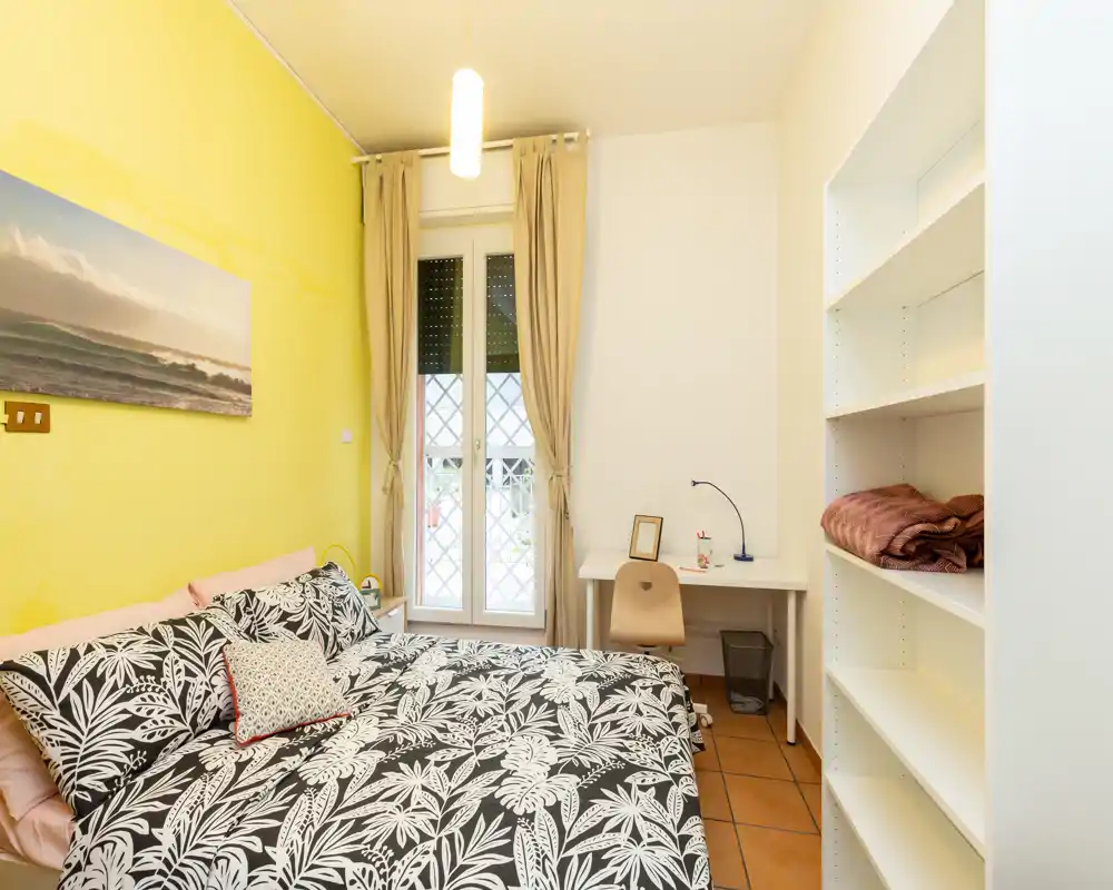 Dormitorio luminoso y bien equipado con cama doble estampada, escritorio junto a la ventana y estanterías empotradas — ideal para una estancia cómoda o estudiantes.