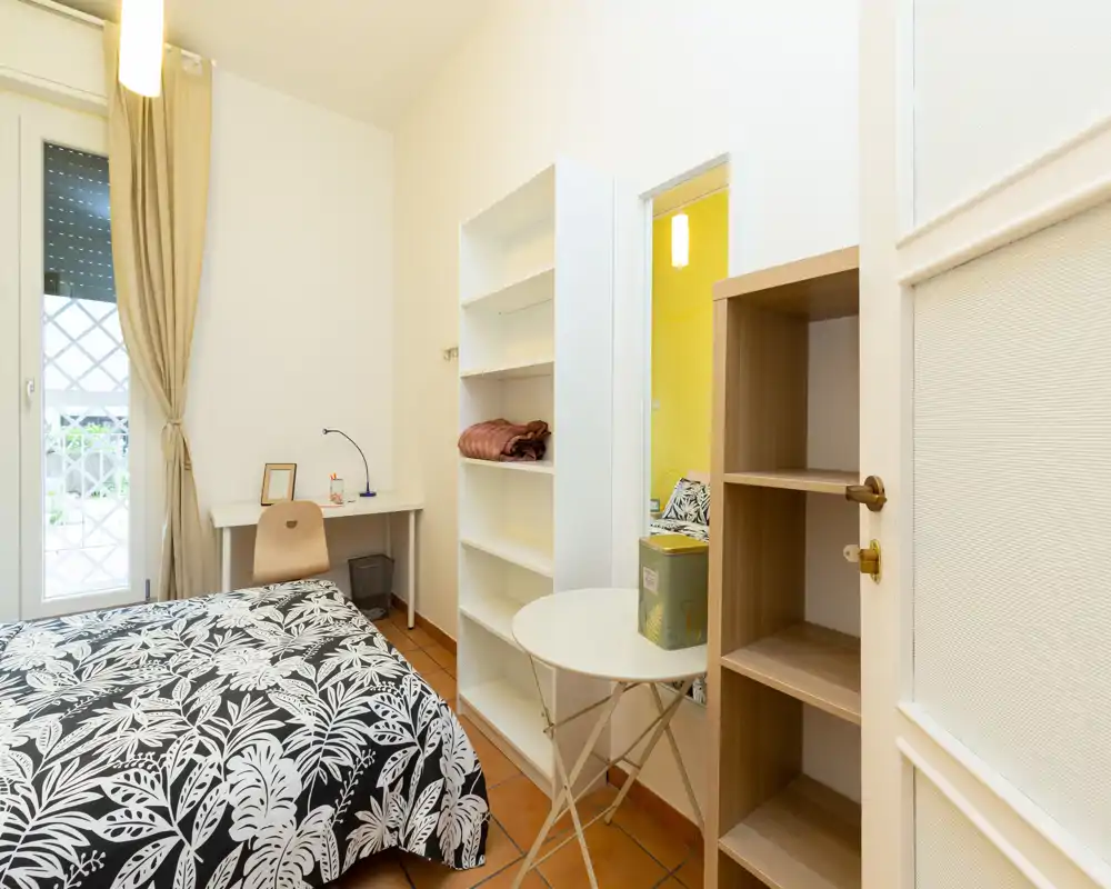 Dormitorio individual luminoso y ordenado con edredón estampado, escritorio junto a la ventana y estanterías abiertas; bien iluminado y amueblado para una estancia confortable.