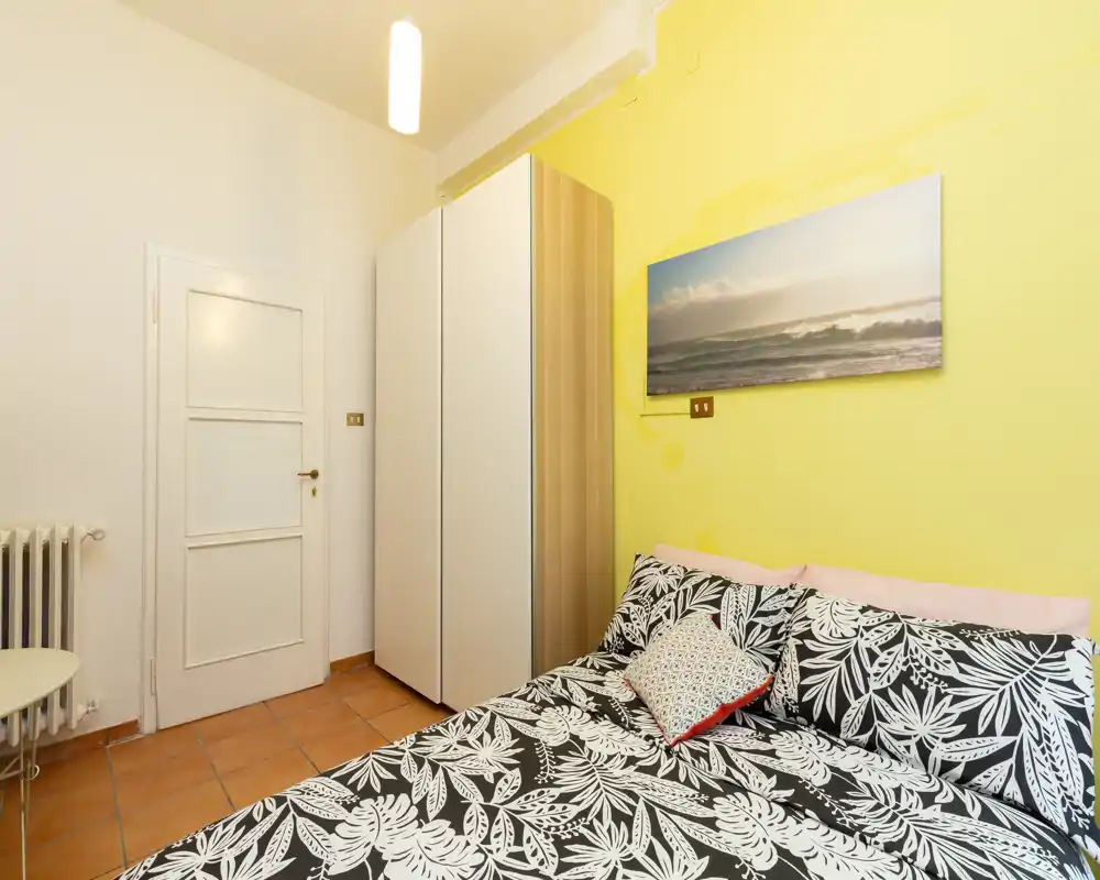 Dormitorio compacto y luminoso con cama doble estampada, armario y pared amarilla cálida — acogedor y perfecto para estancias cortas.