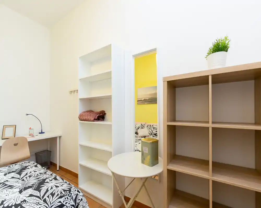 Dormitorio luminoso y ordenado con edredón estampado, escritorio y estanterías; decoración sencilla y moderna con buena luz natural, perfecto para una sola persona.