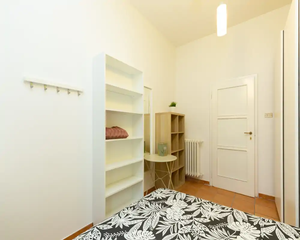 Dormitorio pequeño y ordenado con colcha estampada, estanterías abiertas y un pequeño rincón de escritorio; iluminación cálida y paredes neutras que ofrecen un ambiente tranquilo.