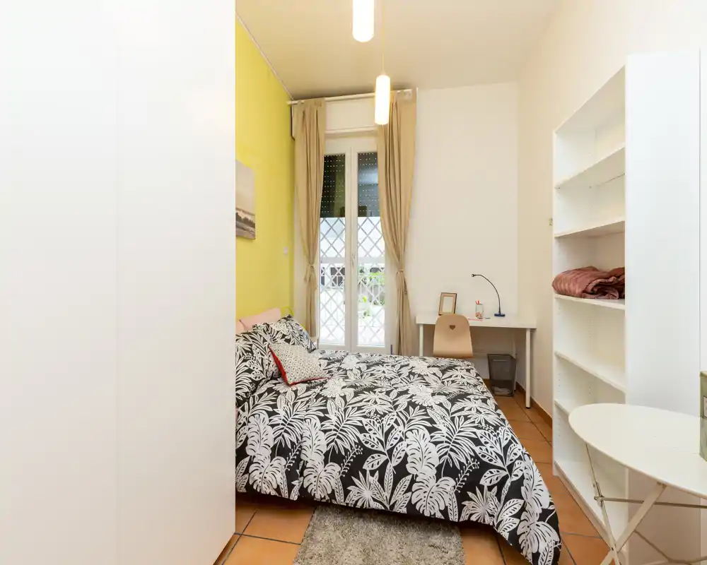 Dormitorio luminoso y ordenado con cama doble cubierta con un edredón de motivos botánicos, un pequeño escritorio junto a la ventana y estanterías abiertas—ideal para estudiante o persona sola.