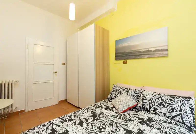 Dormitorio compacto y luminoso con cama doble estampada, armario y pared amarilla cálida — acogedor y perfecto para estancias cortas.